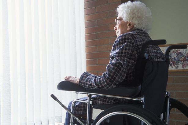 Elderly-woman-in-wheelchair-looking-out-of-window-with-blinds
